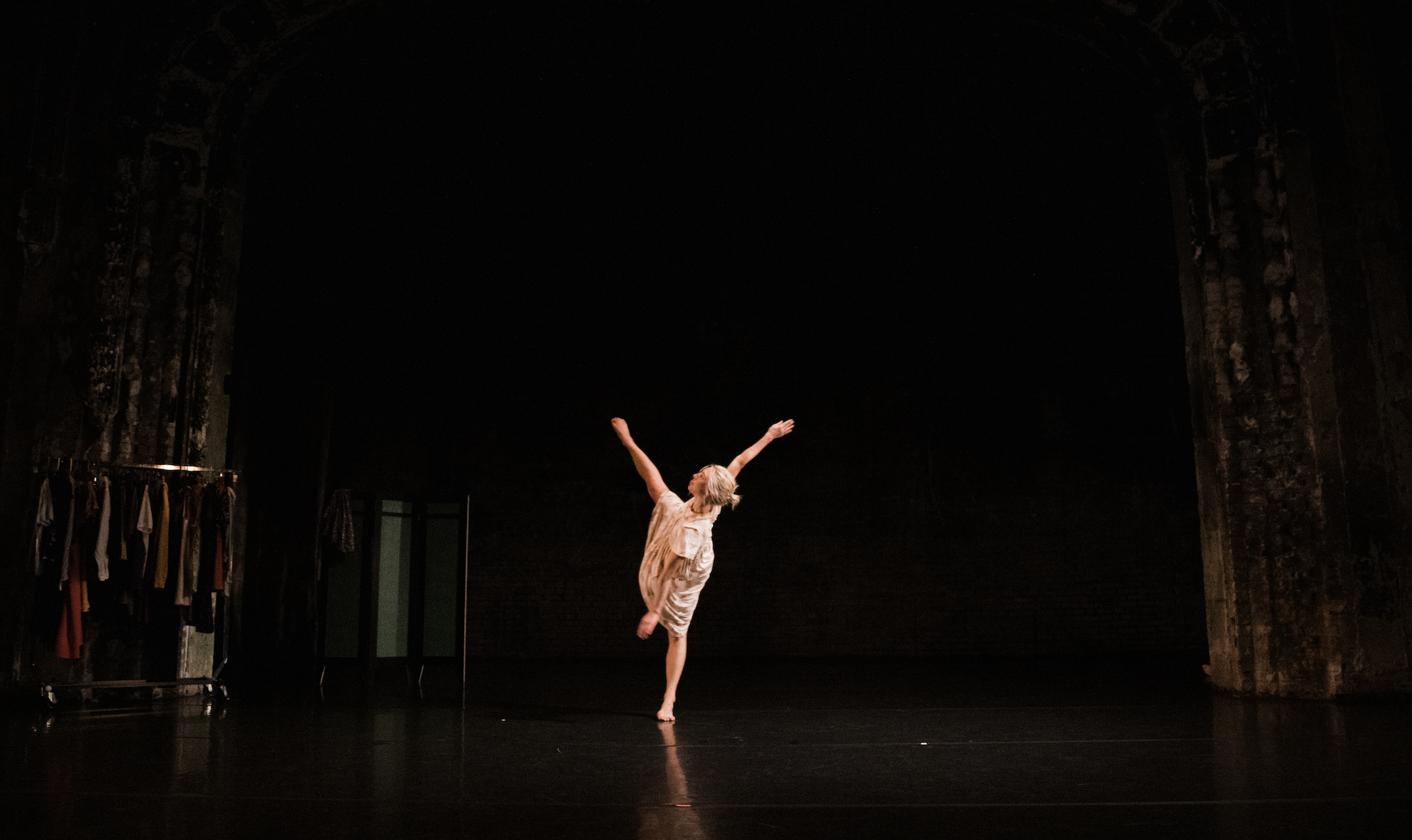 A light-skinned dancer alone on stage in a dress kicks one leg high and tilts to the opposit side
