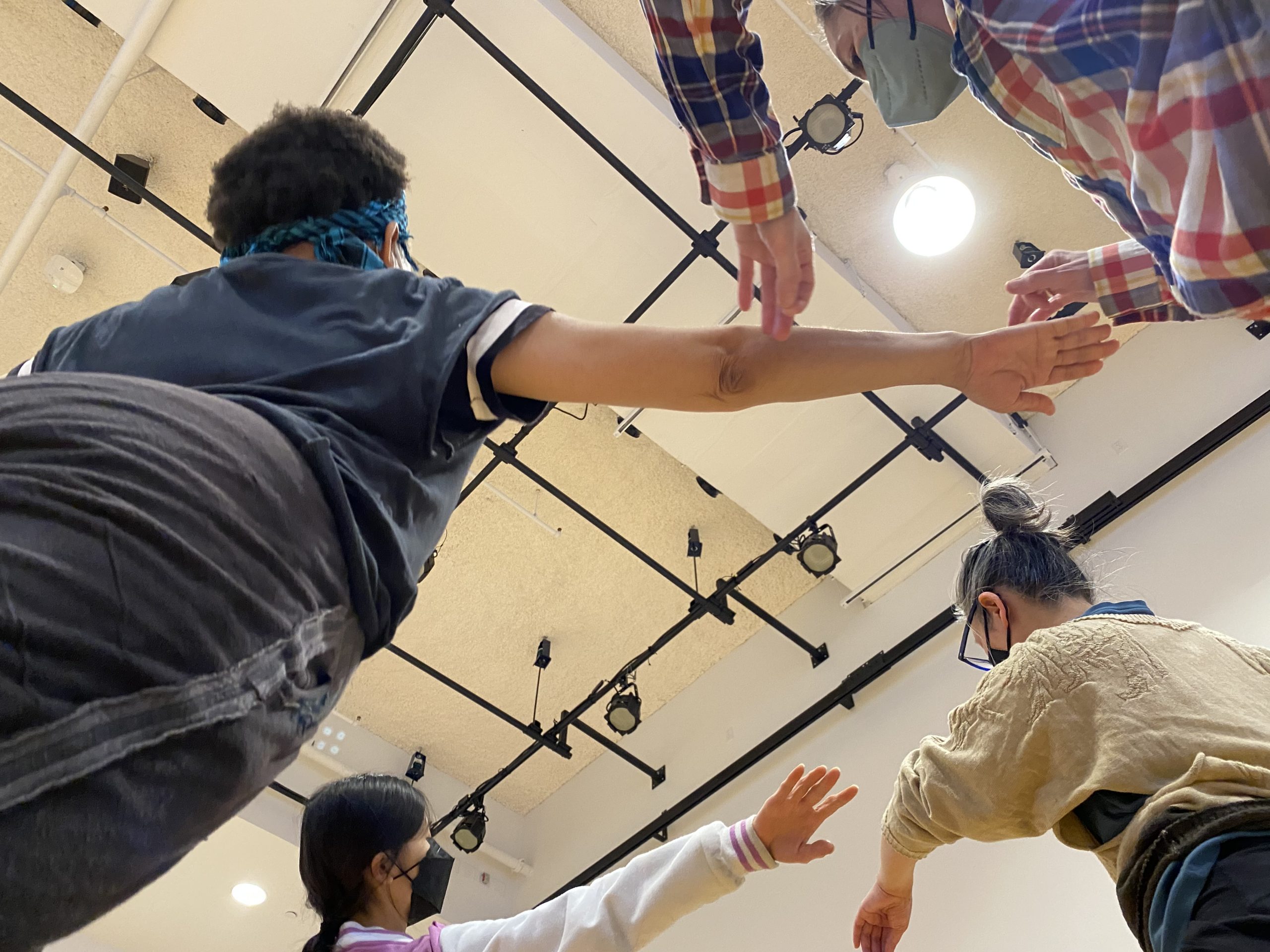A photo taken from a low vantage point, so we see four dancers' outstretched arms and the ceiling of a large space in the background