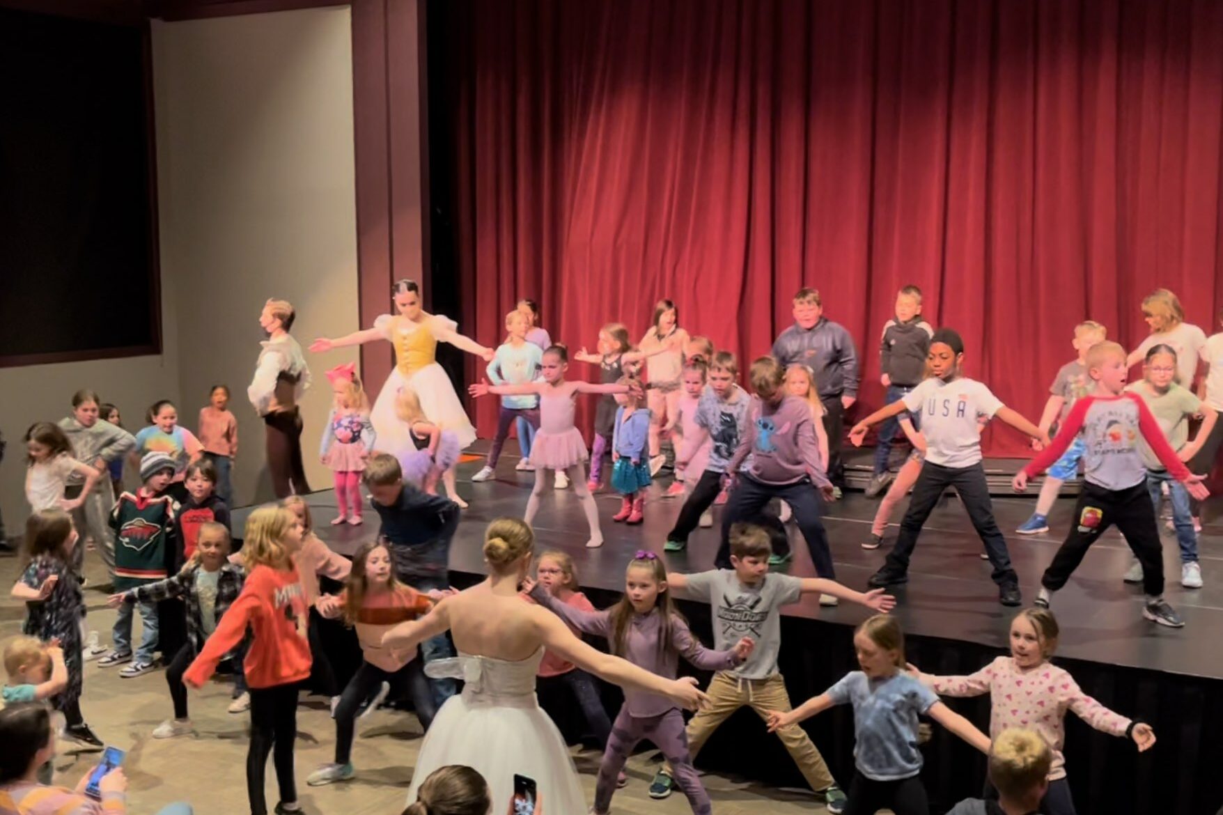 A large group of students of various skin tones and genders stand stand with legs and arms outstretched in a theater