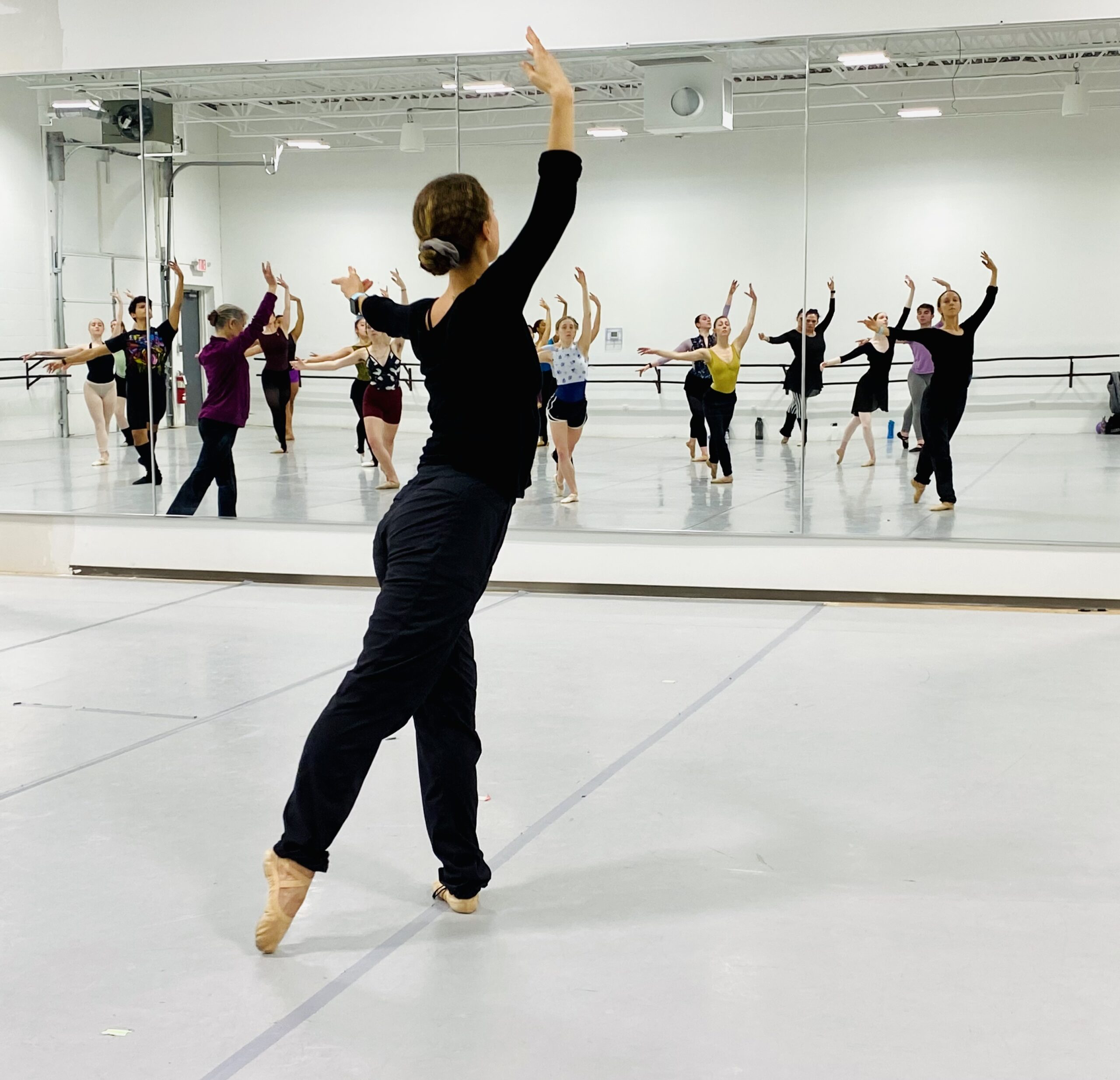 A teacher demonstrates tendu derrier while several students follow in the mirror