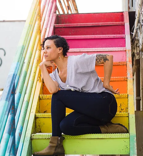 Portrait of Cara Hagen, kneeling with legs intertwined and head resting on chin, on rainbow painted metal stairs